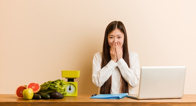 Jonge voedingsdeskundige Chinese vrouw die met haar laptop werkt die om iets lacht, die mond behandelt met handen.