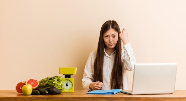 Jonge voedingsdeskundige Chinese vrouw die met haar laptop werkt die vuist toont aan met agressieve gelaatsuitdrukking.