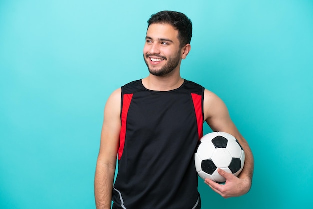 Jonge voetballer Braziliaanse man geïsoleerd op blauwe achtergrond op zoek naar de kant en glimlachen