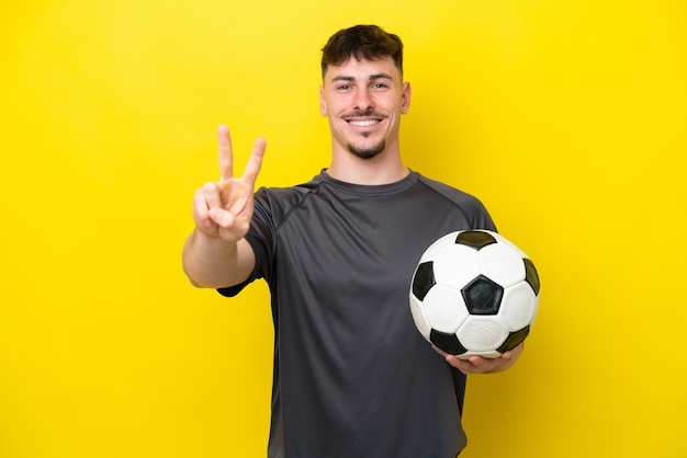 Jonge voetballer man geïsoleerd op gele achtergrond glimlachend en overwinning teken tonen