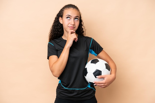 Jonge voetballervrouw die op beige achtergrond wordt geïsoleerd en omhoog kijkt