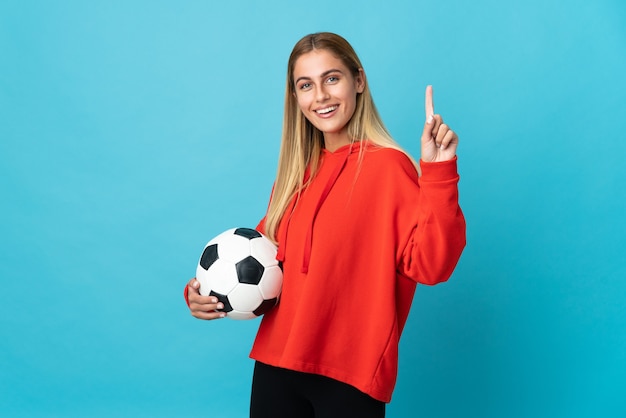 Jonge voetballervrouw die op blauwe achtergrond wordt geïsoleerd die en een vinger in teken van het beste opheft