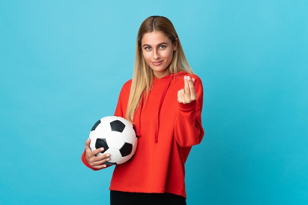 Jonge voetballervrouw die op blauwe muur wordt geïsoleerd die geldgebaar maakt