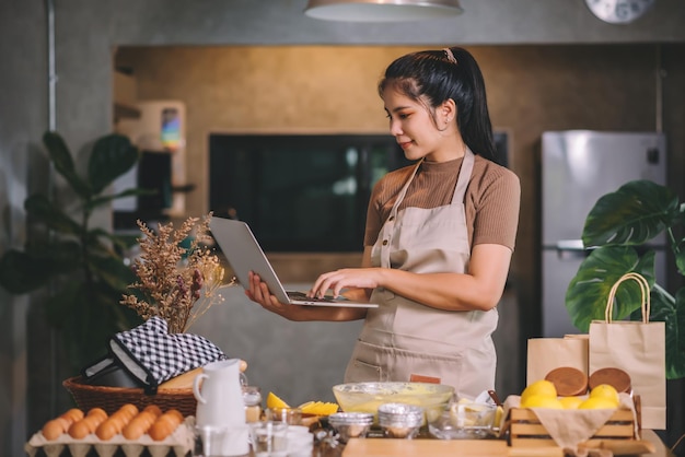 Jonge volwassen aziatische vrouw die zelfgemaakte bakkerij in een keuken thuis voorbereidt en op laptop kijkt naar bakmethoden