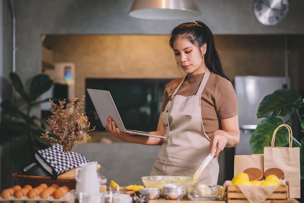 Jonge volwassen aziatische vrouw die zelfgemaakte bakkerij in een keuken thuis voorbereidt en op laptop kijkt naar bakmethoden