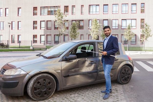 Jonge volwassen bebaarde ongevalsinspecteur inspecteert schadeauto veroorzaakt door auto-ongeluk op de weg autoverzekeringsagent onderzoekt voertuig glimlachend kijkend naar camera met spijkerbroek en jas Buitenschot