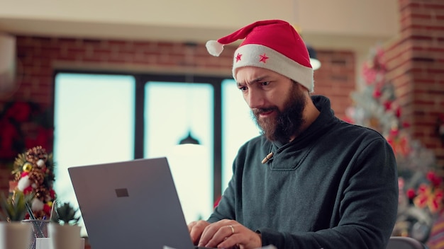 Jonge volwassen brainstormideeën om een rapport op laptop te maken, met een kerstmuts op kantoor met een kerstboom. Feestelijke man aan het werk op computer met onderzoeksgegevens tijdens vakantieseizoen. Uit de hand geschoten.