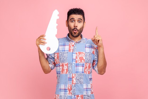 Jonge volwassen man met baard en donker haar, gekleed in een blauw casual stijl shirt met grote papieren sleutel en wijzende vinger omhoog met idee voor het huren van nieuwe flat Indoor studio shot geïsoleerd op roze achtergrond