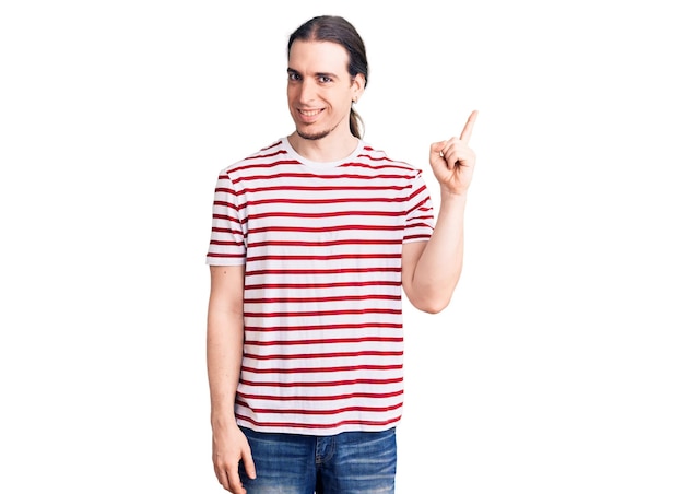 Jonge volwassen man met lang haar, gekleed in casual gestreepte t-shirt glimlachend gelukkig wijzend met hand en vinger naar de zijkant