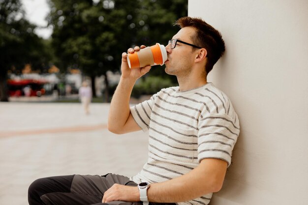 Jonge volwassen man zit buiten koffie te drinken en geniet van mooi weer en uitzicht op de stad Man heeft een pauze van het werk tijd buiten doorbrengen en ontspannen Tijd met jezelf dromen van geestelijke gezondheid