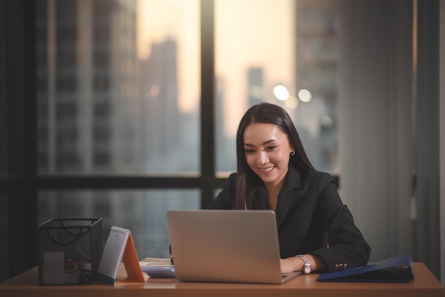Jonge volwassen slimme aziatische zakenvrouw in een zwart, casual pak met behulp van een laptop die overwerkt in een stedelijk kantoor