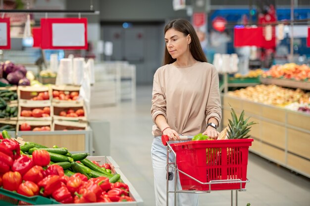 Jonge volwassen vrouw met een casual outfit die door de gangpaden loopt met een winkelwagentje dat dingen kiest om te kopen in een moderne winkel