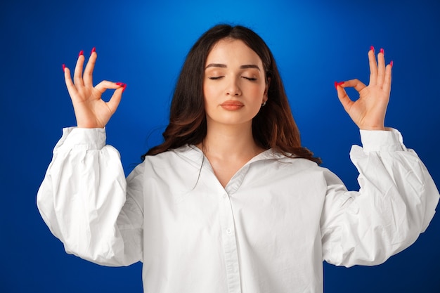Jonge vreedzame vrouw die haar ogen gesloten houdt terwijl ze mediteert tegen een blauwe achtergrond