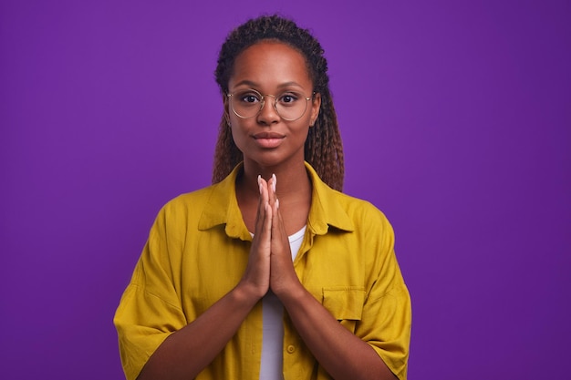 Jonge vriendelijke afro-amerikaanse vrouw vouwt palmen staat in paarse studio