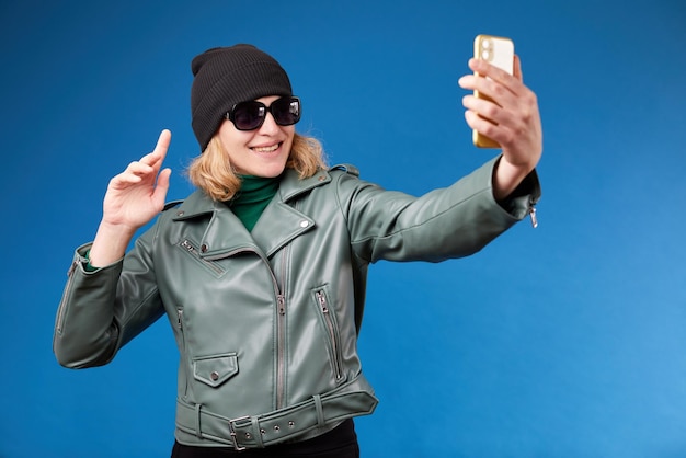 Jonge vriendelijke leuke vrouw 30s dragen stijlvolle groene jas doen selfie geschoten op mobiele telefoon praten door video-oproep zwaaiende hand groet geïsoleerd op blauwe achtergrond studio portret