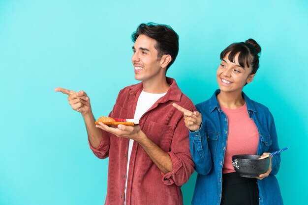 Jonge vrienden van gemengd ras die sashimi en ramen houden geïsoleerd op een blauwe achtergrond wijzende vinger naar de zijkant in zijpositie