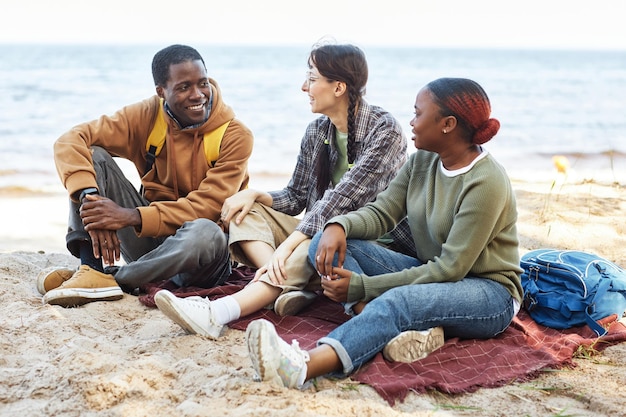 Jonge vrienden zittend op een deken op het zand en praten tijdens picknick in de buurt van de zee
