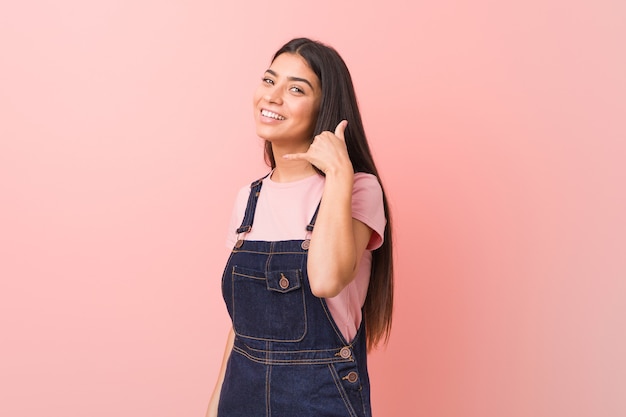 Jonge vrij arabische vrouw die een jeans dungaree draagt die een mobiel telefoongesprekgebaar met vingers toont.