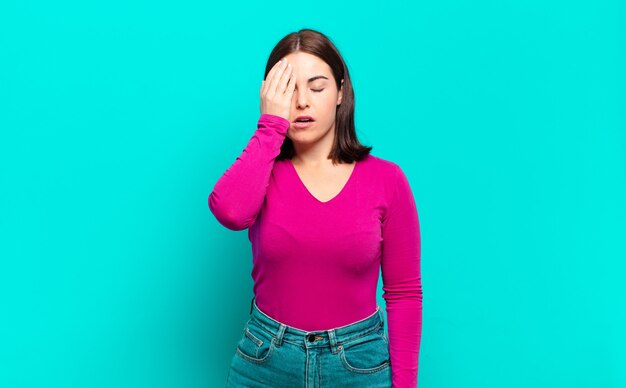 Jonge vrij ongedwongen vrouw die slaperig, verveeld en geeuwend kijkt, met hoofdpijn en één hand die de helft van het gezicht bedekt