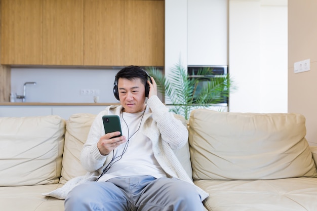 jonge vrolijke aziatische man die naar muziek luistert in een koptelefoon die thuis op de bank zit