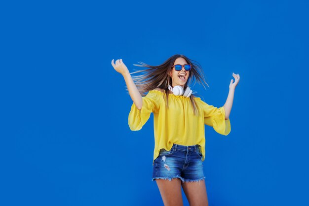 Jonge vrolijke blanke tiener in gele blouse en denim shorts springen voor blauwe muur.