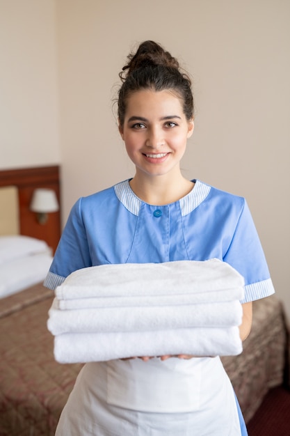 Jonge vrolijke brunette kamer meid met stapel witte schone zachte handdoeken permanent in hotelkamer