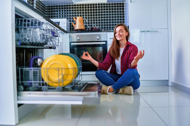 Jonge vrolijke gelukkige huisvrouwenvrouw die moderne vaatwasser gebruikt voor afwas en glazen in de witte huiskeuken. Ontspan en geniet tijdens de schoonmaakklusjes