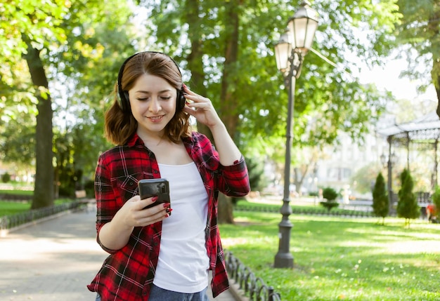 Jonge vrolijke grappige vrouw luistert naar muziek met een koptelefoon en gebruikt smartphone in stadspark
