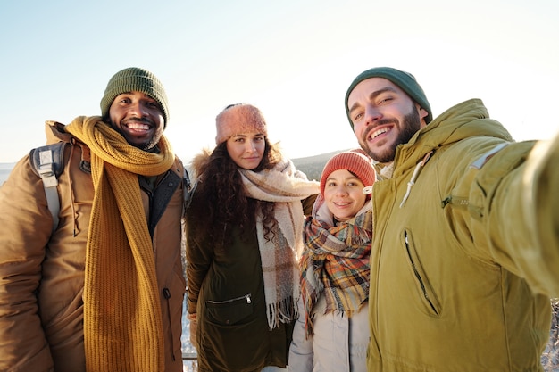 Jonge vrolijke interculturele vrienden in winterkleding die selfie maken voor de camera tegen heldere hemel op een zonnige winterdag terwijl ze aan het chillen zijn