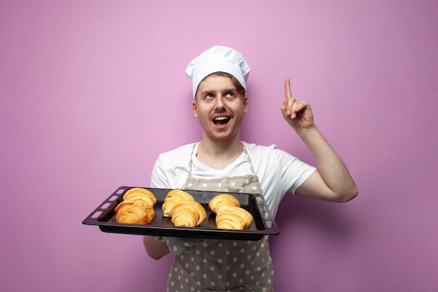 Jonge vrolijke man huisvrouw in een schort en hoed houdt bakplaat met gebakken croissants