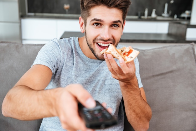 Jonge vrolijke man met afstandsbediening en op de knop te drukken tijdens het eten van pizza.