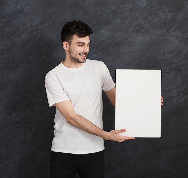 Jonge vrolijke man met lege witte banner en aanbevelen. Glimlachende man met reclameblad en duim omhoog gebaar, grijze studio achtergrond, kopieer ruimte