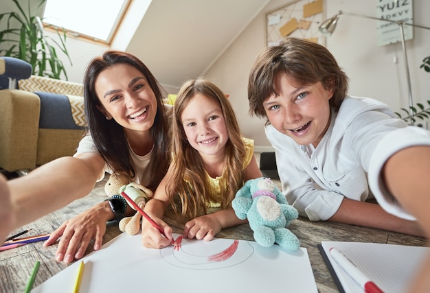 Jonge vrolijke moeder met twee gelukkige kinderen die op de vloer in de woonkamer liggen en maken