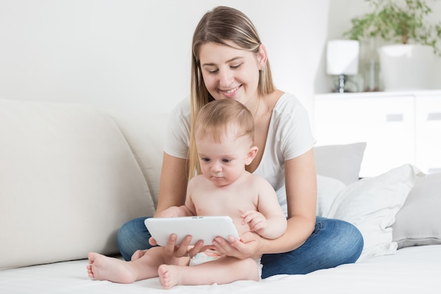 Jonge vrolijke moeder zittend op de bank met haar babyjongen en surfen op internet op tablet-pc