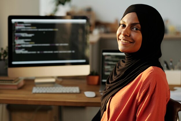 Jonge vrolijke moslim vrouwelijke werknemer in hijab en rood shirt die naar je kijkt