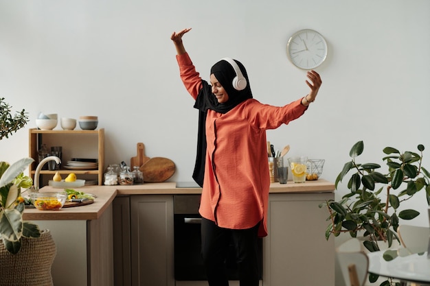 Jonge vrolijke moslimvrouw in hijab en koptelefoon die in de keuken danst