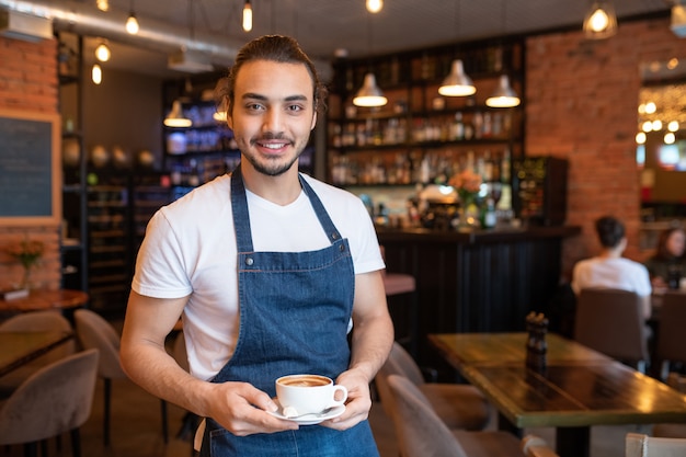 Jonge vrolijke ober in wit t-shirt en blauw schort kopje cappuccino op schotel te houden terwijl je naar je kijkt