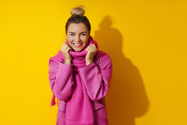 Jonge vrolijke vrouw die roze jas en wollen sjaal draagt tegen gele achtergrond