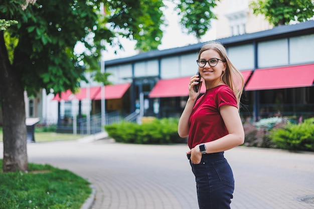 Jonge vrolijke vrouw in grijze trui en spijkerbroek en bril die op de stoep staat met boodschappentassen en berichten op smartphone in New York