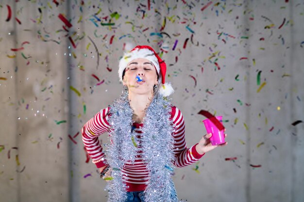 jonge, vrolijke zakenvrouw die een rode hoed draagt en een feestfluitje blaast terwijl ze een nieuwjaarsconfetti-feestje heeft voor een betonnen muur