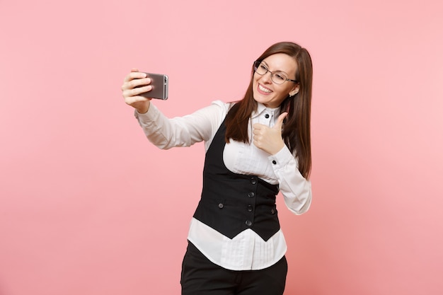 Jonge vrolijke zakenvrouw in pak, bril doet selfie schot op mobiele telefoon met duim omhoog geïsoleerd op roze achtergrond. Dame baas. Prestatie carrière rijkdom. Kopieer ruimte voor advertentie.