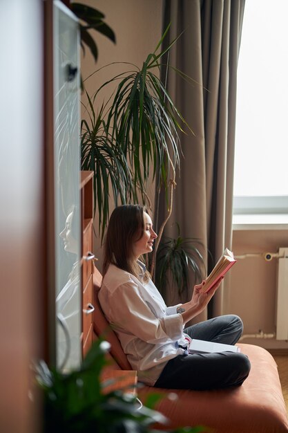 Jonge vrolijke zelfverzekerde en gelukkige vrouw ontspant thuis in haar gezellige fauteuil met boeken