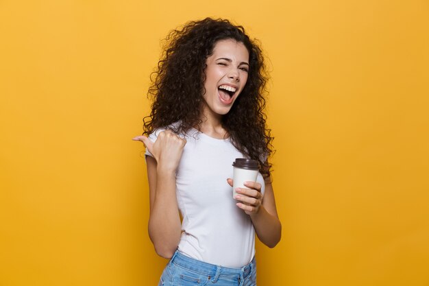 jonge vrouw 20s met krullend haar glimlachend en met afhaalmaaltijden koffie in papieren beker geïsoleerd op geel