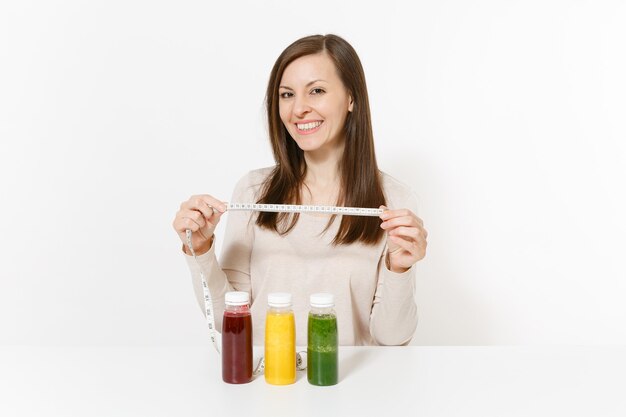 Jonge vrouw aan tafel met groene, rode, gele detox smoothies in flessen, meetlint geïsoleerd op een witte achtergrond. goede voeding, vegetarische drank, gezonde levensstijl, dieetconcept. ruimte kopiëren.