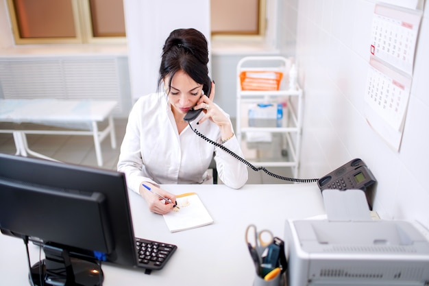 Foto jonge vrouw arts die telefonisch spreekt en iets in haar bureau schrijft