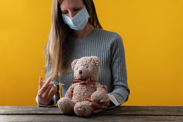 Jonge vrouw, arts, met medisch beschermend masker die een teddybeer vaccineert. Over gele achtergrond.