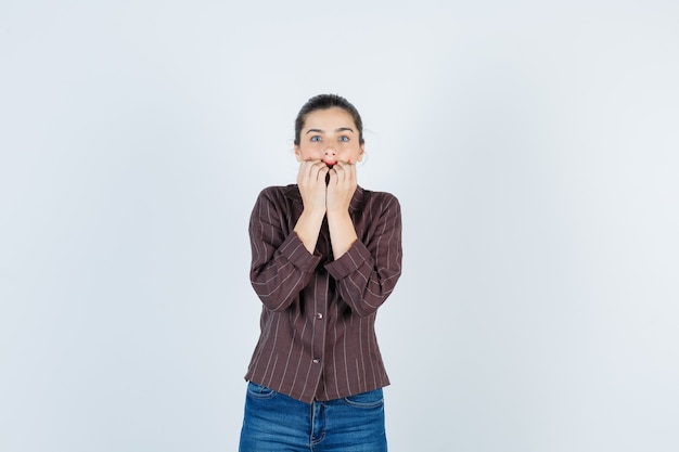 Jonge vrouw bijt emotioneel vuisten in gestreept shirt, spijkerbroek en kijkt angstig, vooraanzicht.