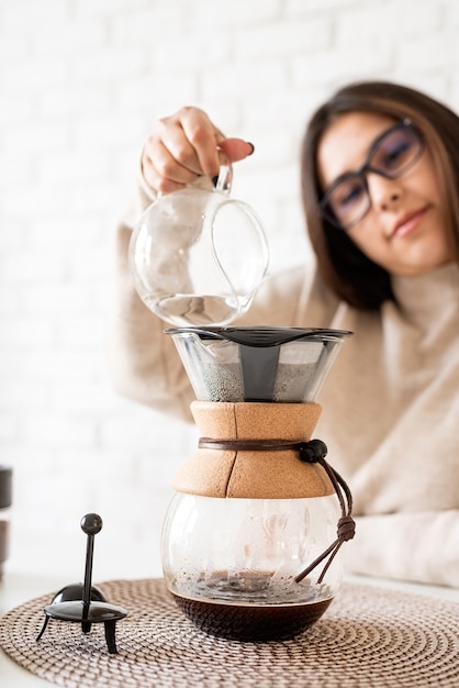 Jonge vrouw brouwen van koffie in coffemaker staande aan de witte tafel met verschillende spullen voor alternatieve koffie brouwen, heet water gieten in het filter