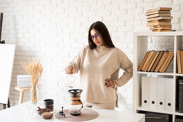 Jonge vrouw brouwen van koffie in coffemaker staande aan de witte tafel met verschillende spullen voor alternatieve koffie brouwen, heet water gieten in het filter