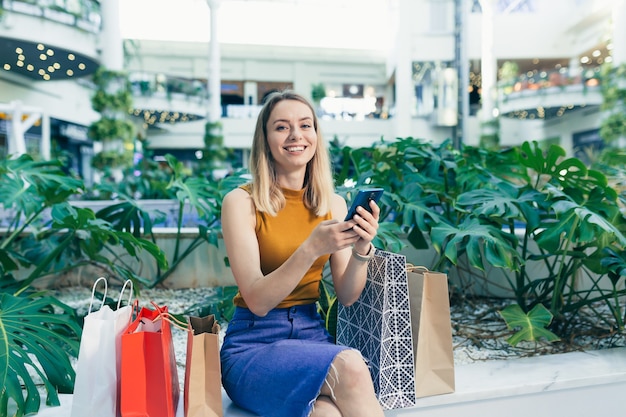 Jonge vrouw consument in het winkelcentrum bladert door chat en gebruikt het gebruik van een smartphone. vrouw staat met een mobiele telefoon in haar handen in het winkelcentrum. binnen. gelukkig shopper meisje met cadeauzakjes aankopen doen